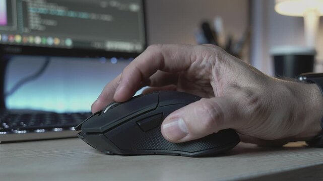 Close-up of a male hand using a computer mouse on a wooden desk. The background features a blurred monitor screen, suitable for technology, business, work, and gaming concepts.