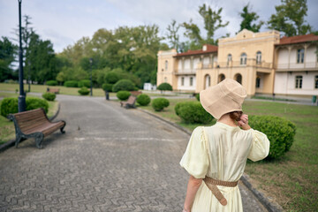 Obraz premium Woman in a vintage dress and straw hat walks along a park path toward a grand estate, creating a serene outdoor scene with timeless charm today.