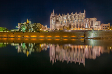 Night view of Palma Cathedral in Mallorca, beautifully illuminated against the dark sky, reflecting in calm waters and showcasing the stunning Gothic architecture of this iconic landmark.