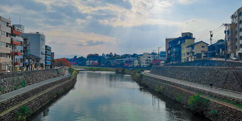 Fototapeta premium Serene river meanders through a traditional Japanese town, its calm waters reflecting charming buildings under a dramatic, cloudy sky, evoking peaceful urban beauty and local culture.