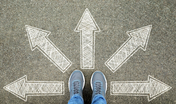 Man in sneakers standing on road with arrows pointing in different directions, top view. Start your journey