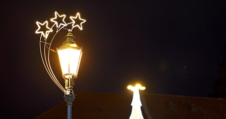 Illuminated lamp stands tall. Bright stars adorn the fixture. A festive glow emanates. Dark background enhances the light. Architectural detail is visible. Night scene creates ambiance © rikajiMiAda