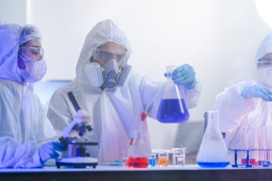 Team of professional Asian scientists wearing protective hazmat suits analyzing blue chemical liquid in modern biotechnology laboratory research safety industrial development
