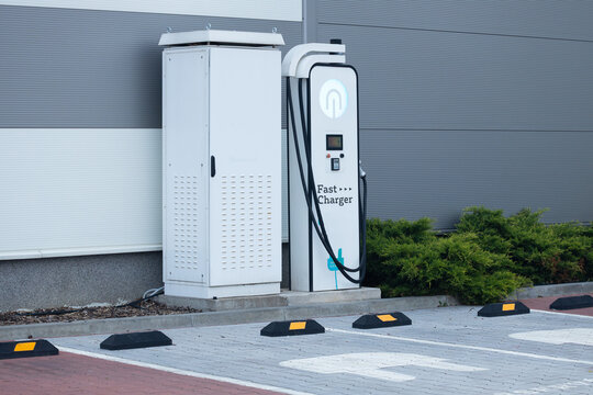 Electric vehicle fast charger station with charging unit and power supply cabinet located in a parking area beside green shrubs and gray building wall