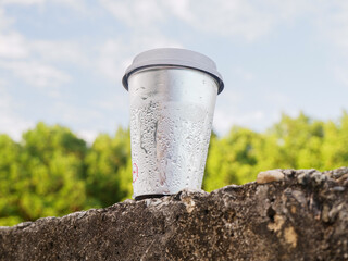 ​Silver metallic beverage cup mockup with condensation droplets on a stone wall. Blurred nature...