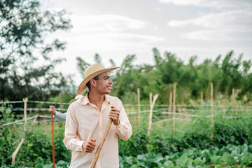 Happy young male farmer in a straw hat holding a garden tool and looking away while standing in an organic vegetable field.