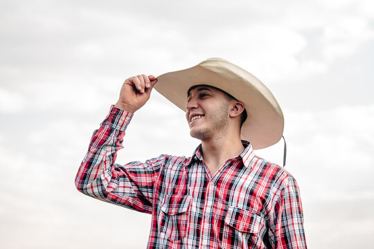 Happy young cowboy in a plaid shirt and straw hat smiling outdoors against a bright sky