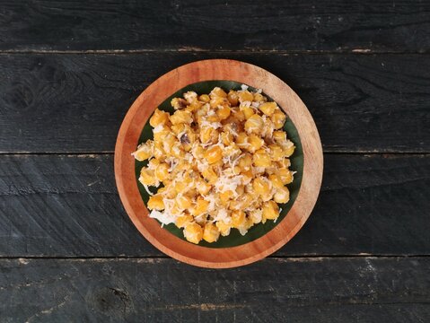 Top view of urab jagung, a traditional indonesian sweet snack made from boiled corn kernels mixed with grated coconut and sugar. Served on wooden plate with black wooden table