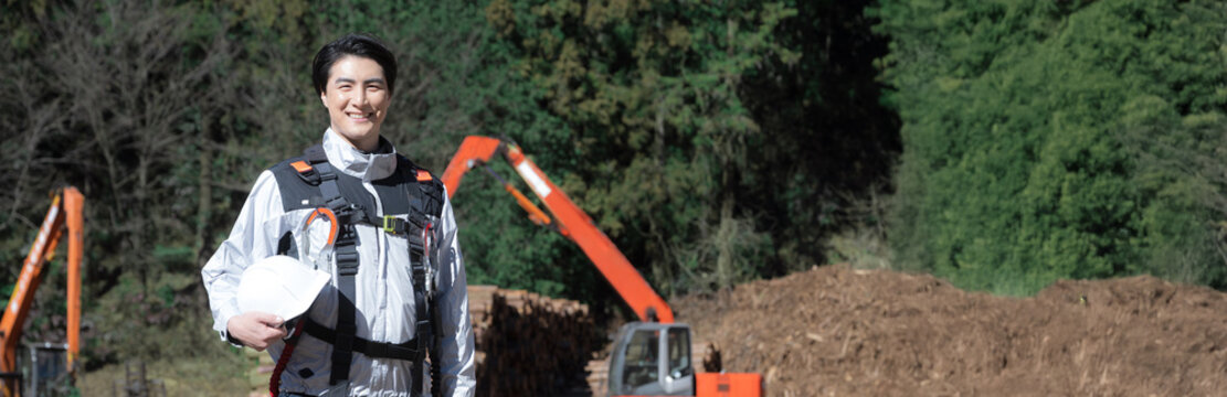 建築現場などでショベルカーを背景にヘルメットをつけた林業など高所作業などのハーネス(安全帯)を付けた男性カメラ目線の笑顔　春夏の緑背景　(多摩木材センター協同組合プロパティ許可ずみ）バナー