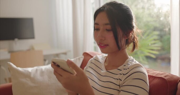 Asian teen texting, message, or chat on smartphone. Late afternoon light filters through trees in living room