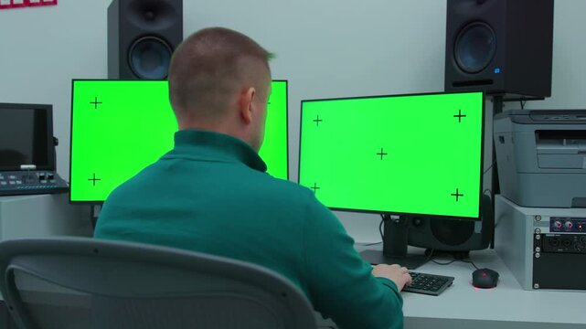 A man sitting on a working chair and working in the scene with two computer monitors on a desk with a green screen.