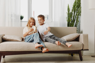 Two children sitting barefoot on beige sofa at home, looking at smartphone together in bright...