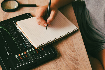 Businessman writing financial strategy in notebook with stock market charts on tablet, concept of investment planning and data analysis.
