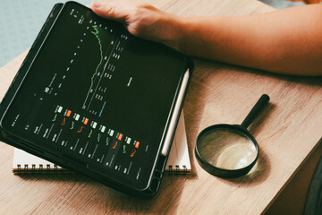 Close up of businessman holding tablet with stock market charts, analyzing financial data with magnifying glass on wooden desk.
