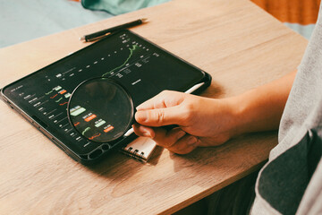 Close up of hand holding magnifying glass analyzing stock market data on tablet, concept of financial audit and investment research.
