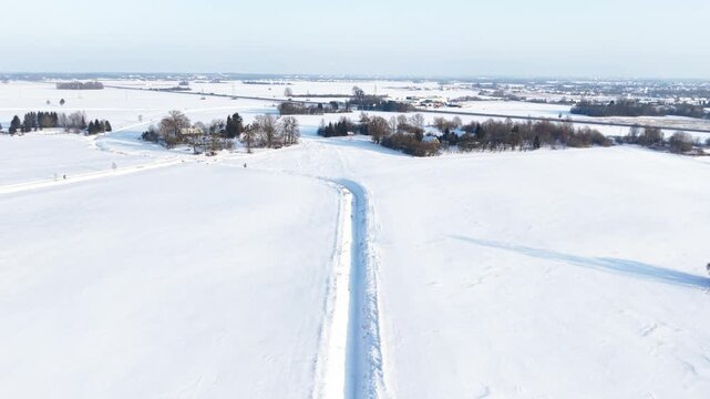 Drone video capturing a serene, snow-blanketed rural landscape under a clear blue sky. The expansive fields, dotted with trees and distant buildings, evoke a peaceful winter mood.