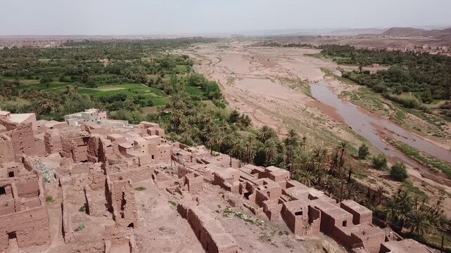 Aerial shot of ancient Kasbah ruins alongside a lush river oasis. The location highlights historical human settlement patterns and adaptation to water resources in the arid Moroccan desert.