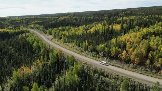 Tracking aerial shot of a white van driving along a curving unpaved Dempster Highway in Canada, surrounded by mixed forest with vibrant green and yellow foliage in early autumn.