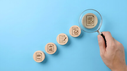 Fototapeta premium Hand,holding,magnifying,glass,over,wooden,blocks,showing,step,by,step,document,approval,process,icons,business,compliance,and,quality,control,concept,on,blue,background,with,checked,files.