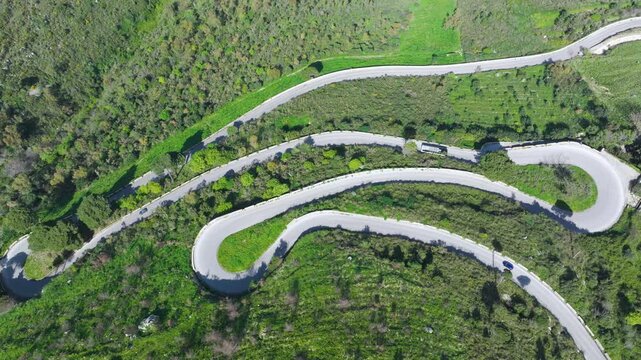 AERIAL TOP DOWN: Narrow mountain road carving tight hairpin turns through hillside covered with lush green Mediterranean greenery. Vehicles overcome serpentines through countryside in western Sicily.