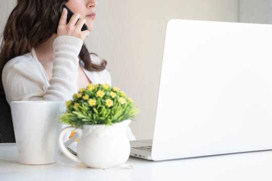 Mujer atendiendo llamada telef&oacute;nica en su oficina en casa con laptop, taza de caf&eacute; y maceta decorativa.