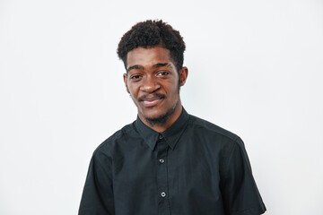 Obraz premium Young black man with dark skin wearing black shirt standing against white background looking at camera with slight smile and relaxed posture in studio portrait.