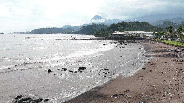 A scenic aerial perspective of the Limbe coastline, featuring a mix of residential houses, dark volcanic sand, and the lush foothills of Mount Cameroon