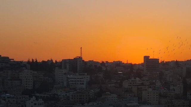 Stunning sunset over Amman, Jordan, as the sun disappears behind hillside neighborhoods, bathing the city skyline in orange light while a flock of birds crosses the evening sky