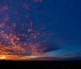 Obraz premium A wide view of a dramatic sunset sky filled with dense textured clouds glowing orange and blue, stretching above a dark distant horizon and creating a strong natural background atmosphere