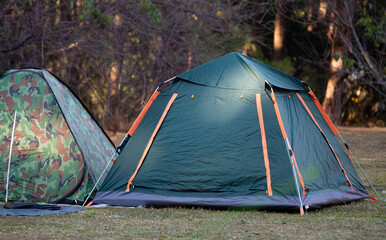 Camping Concept: Dark Green Outdoor Tent and Camouflage Pop-Up Tent Set Up on Grass in the Forest with Natural Sunlight During a Summer Trip, Adventure Leisure Activity and Recreation Background.