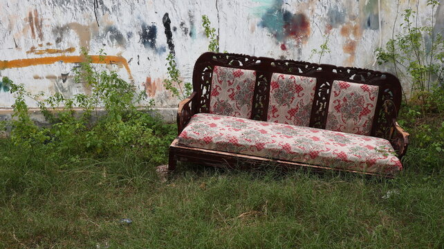 Worn wooden couch abandoned in grass near a cracked graffiti wall, symbolizing neglect, decay, and forgotten urban environments.