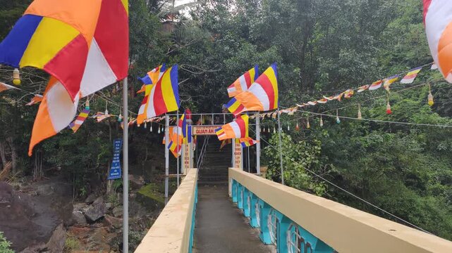 Wide view shows Sacred Pathway with colorful Buddhist flags waving on bridge. Tropical forest surrounds entrance to temple during Sacred Pathway journey in mountains.