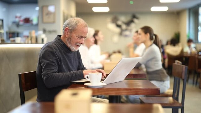 Senior man is working on a laptop in a cozy cafe, drinking coffee and typing intently, he is doing remote work. Elderly writer types a new chapter for a book. High quality 4k footage
