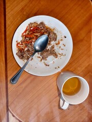 Leftovers of a meal with buckwheat and meat on a white plate with a spoon, and a cup of tea on a wooden table.