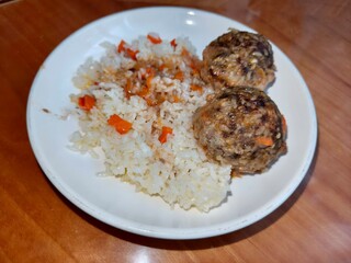 Plate served with two meatballs with rice and garnished with white rice and diced carrot. Homemade dinner with hearty food menu item.