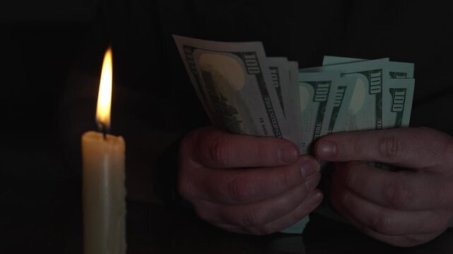 Man's hands counting a stack of hundred-dollar bills in a dark, mysterious room illuminated only by the warm, flickering flame of a single candle, creating a secretive atmosphere