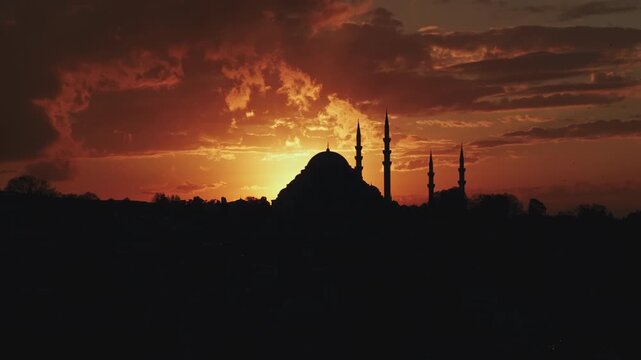 A cinematic aerial drone shot of Suleymaniye Mosque in Istanbul during sunset, with the sun setting directly behind the mosque. The warm golden light creates a dramatic silhouette of the historic Otto