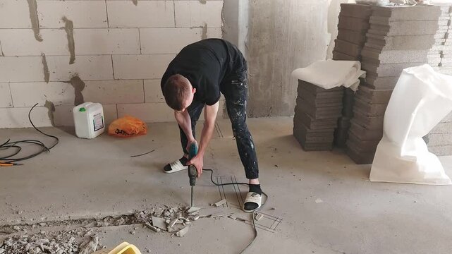 A man drills into the floor in a building site. Concrete blocks are stacked nearby, and tools are spread out around the area. The work continues in the construction zone.