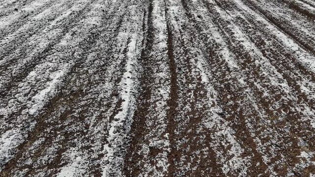Stunning aerial drone perspective of a snow-covered ploughed field. The deep contrasting parallel lines of dark brown soil and bright white snow create a striking geometric pattern.