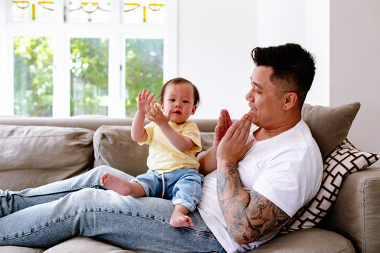 Asian father and baby girl clapping while seated on the couch