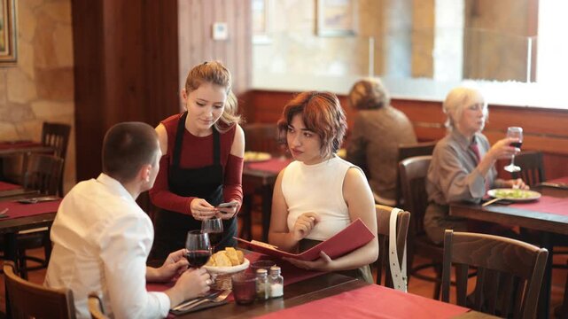 Polite female server helping young couple with choice in warmly decorated restaurant environment