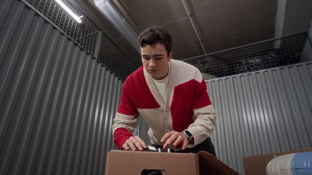 Young man opening cardboard box, retrieving pair of boxing gloves and golden trophy, examining his belongings and smiling inside personal storage unit