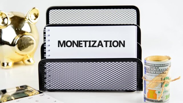 The word monetization gradually forms on a white notebook. A golden piggy bank sits nearby with a stack of US hundred dollar bills