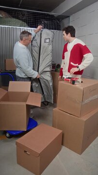 Vertical of middle aged man and his teenage son unpacking brown winter jacket from garment bag and discussing it, surrounded by labeled cardboard boxes in storage unit