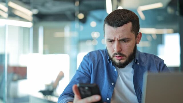 Man experiences range of emotions from surprise to concern and disappointment while viewing content on smartphone in modern office environment, laptop nearby.