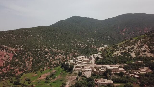 Aerial wide shot of a remote Berber village situated on the rugged red slopes of the High Atlas Mountains. The footage highlights the geological textures, red earth, and rural settlements in North Afr