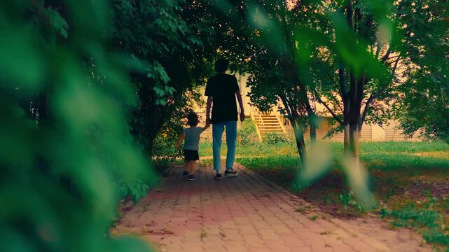 Father and young son walking hand in hand through tree archway in summer park