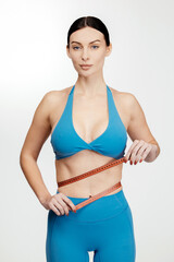 Fototapeta premium Female model measuring waist with tape measure, wearing blue sports bra and leggings, standing against a plain white background, promoting body shape control and healthy lifestyle