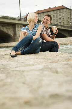 Young couple sitting by riverfront enjoying takeaway coffee