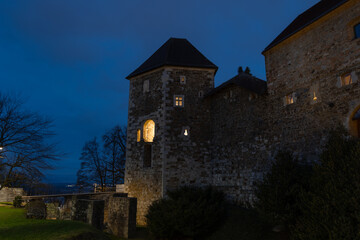 Fototapeta premium Twilight by Ljubljana Castle in Slovenia with glowing windows and stone walls, bare winter trees and deep blue sky, photographed in January from the hilltop fortifications